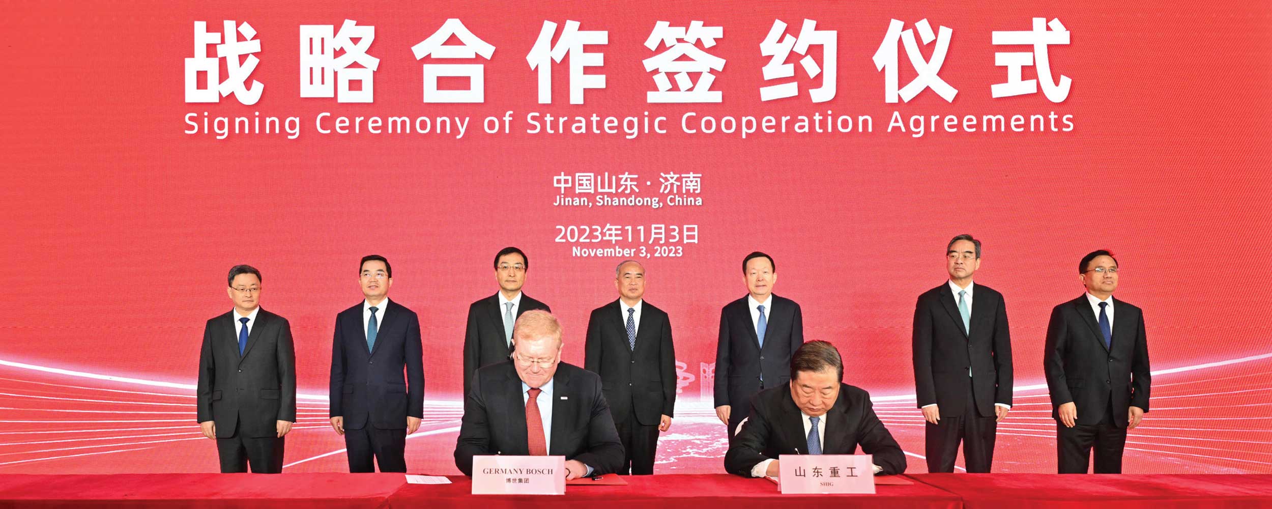 Seven men stood in front of a red background with two men sat at a table.